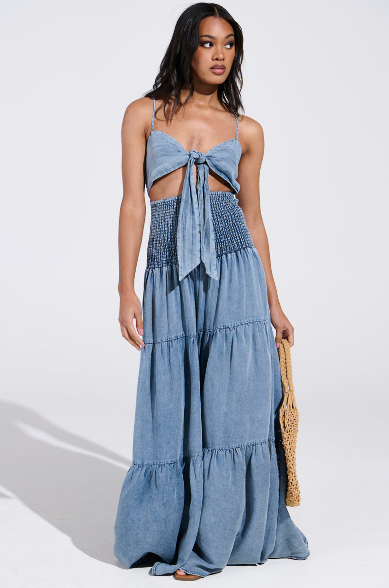 In a studio, a woman stands wearing the JUMPIN AROUND JUMPSUIT. The denim piece features adjustable tank straps, a bow with a front cutout, and exudes effortless charm with its smocked waist and tiered skirt. She holds a woven bag in her left hand and gazes to the side.