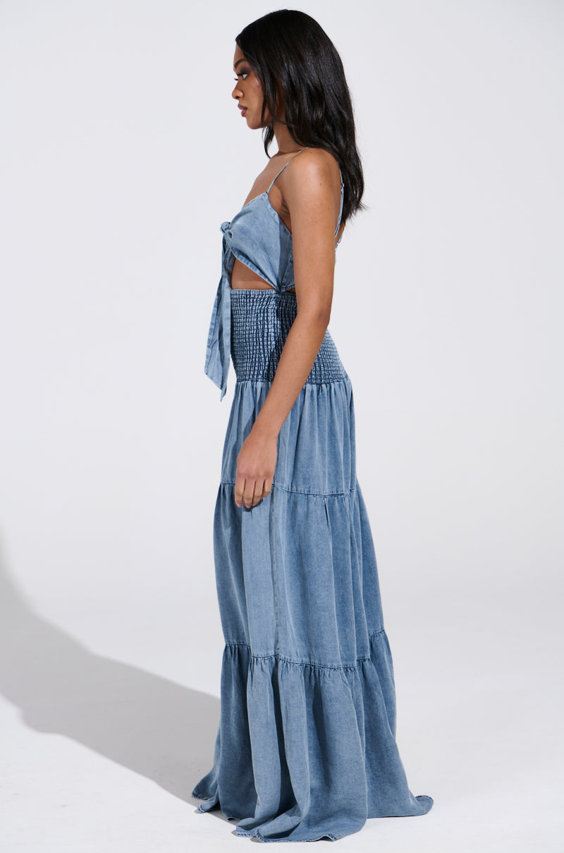 Someone in a sleeveless, JUMPIN AROUND JUMPSUIT with adjustable straps and a tiered skirt stands in profile. Their smocked denim attire complements their long dark hair against a plain background.