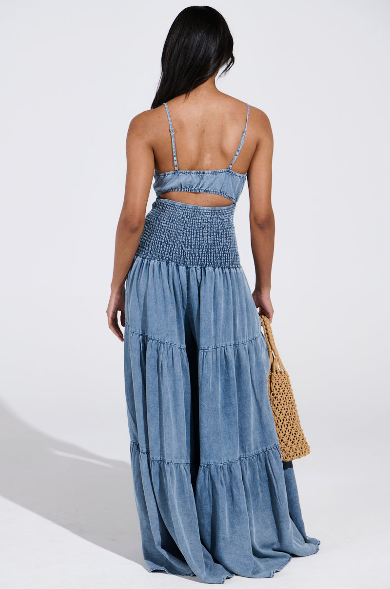 A person with long dark hair, wearing the JUMPIN AROUND JUMPSUIT—a blue, sleeveless outfit with adjustable tank straps and a smocked waist—stands facing away. They hold a woven bag in their left hand against a plain, well-lit background.