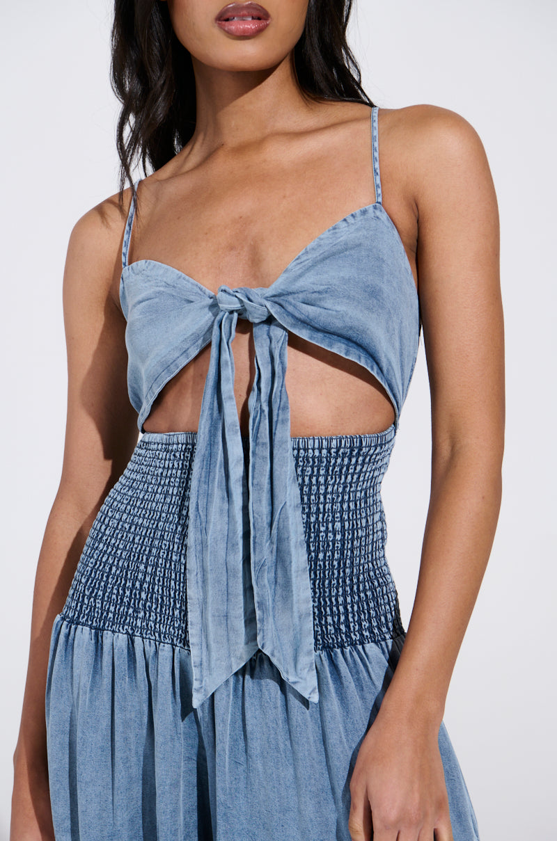 A woman wears the JUMPIN AROUND JUMPSUIT, a blue denim piece with adjustable tank straps and front tie detail. It features a smocked waist and flowy skirt, resembling a stylish denim jumpsuit against a plain background that highlights its elegant vibe.