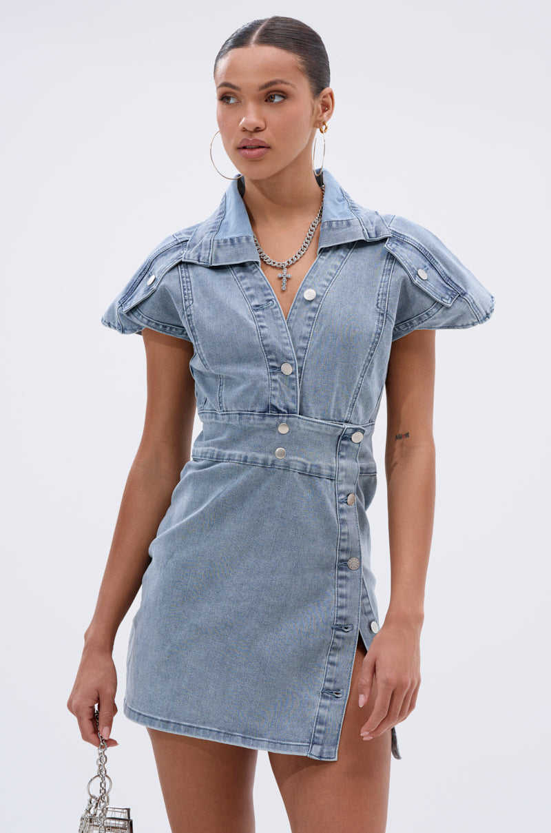 A woman stands against a plain background wearing the NEVER TOO MUCH DENIM MINI DRESS, featuring a collar and front slit. She holds a small handbag and pairs hoop earrings with layered necklaces—ideal for day-to-night looks.