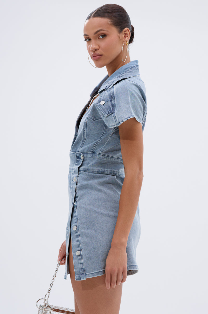 A woman stands sideways against a white background wearing the NEVER TOO MUCH DENIM MINI DRESS—a short-sleeved, collared, button-up mini dress with front pockets. She holds a small handbag, wears large hoops, and has her hair pulled back.