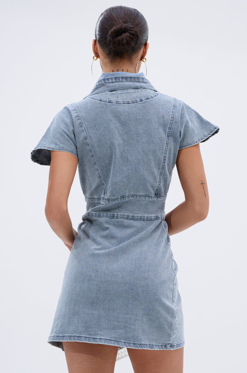 A woman with her hair in a bun wears the NEVER TOO MUCH DENIM MINI DRESS, featuring short sleeves and a belt. She stands with her back to the camera, showing a small cross tattoo on her left arm—ideal for day-to-night wear.