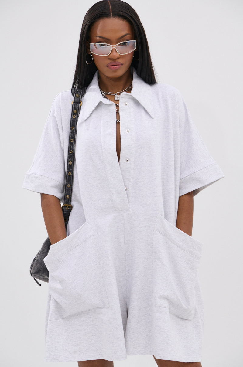 A woman wears the SHE'S RICH OVERSIZED FRENCH TERRY ROMPER in light gray with large front pockets, styled with a silver chain necklace, hoop earrings, clear cat-eye glasses, and a black shoulder bag for all-day comfort and effortless luxury.