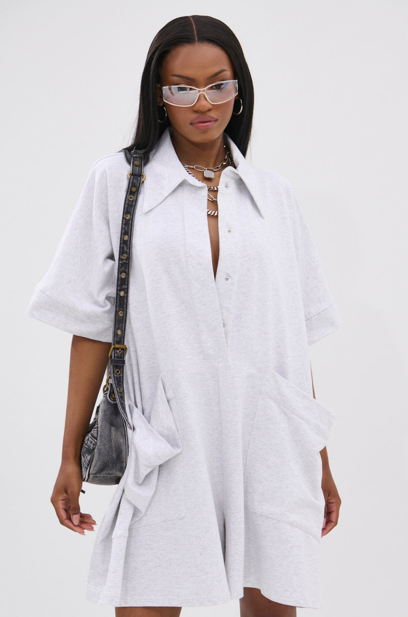 Wearing the SHE'S RICH OVERSIZED FRENCH TERRY ROMPER with large front pockets, layered necklaces, clear cat-eye sunglasses, and a black shoulder bag, she exudes effortless luxury and comfort against a plain white background.