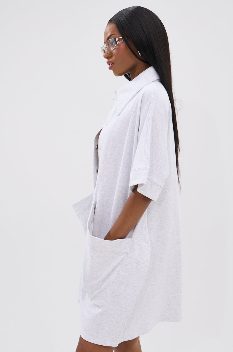 A woman with long straight hair and glasses models the SHE'S RICH OVERSIZED FRENCH TERRY ROMPER, a loose, light gray, short-sleeve romper with pockets—ideal for all-day comfort and effortless style against a white backdrop.