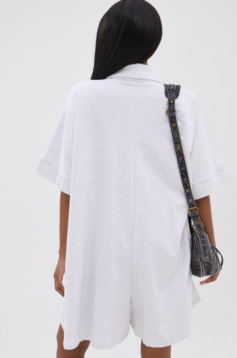 Seen from the back, a person with long, straight dark hair wears the SHE'S RICH OVERSIZED FRENCH TERRY ROMPER in light gray for all-day comfort, paired with a black shoulder bag with gold hardware against a plain light background.