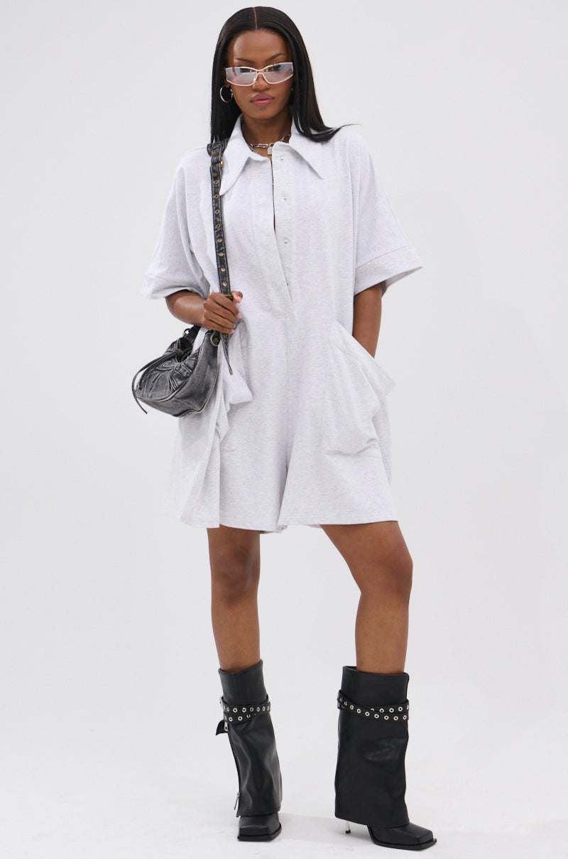 A woman in the SHE'S RICH OVERSIZED FRENCH TERRY ROMPER with large pockets, black studded knee-high boots, sunglasses, and a metallic shoulder bag stands against a plain white background.