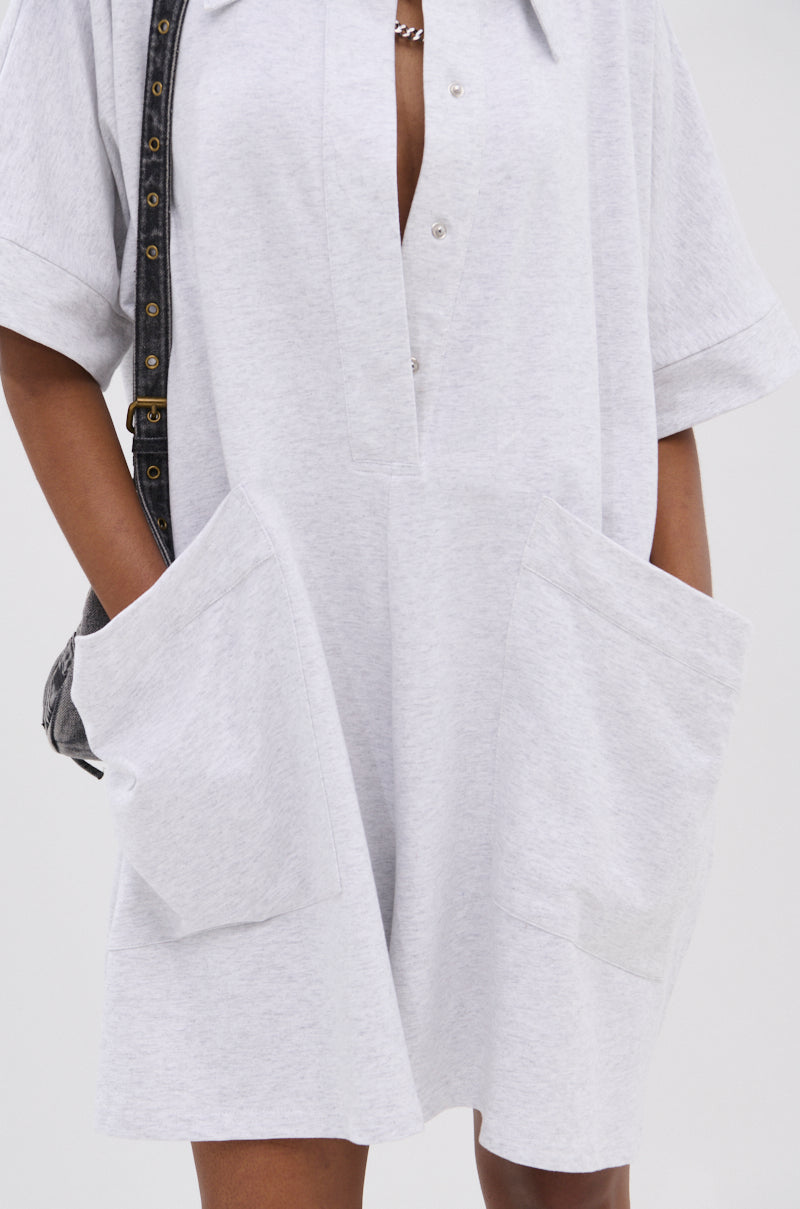 A person wears the SHE'S RICH OVERSIZED FRENCH TERRY ROMPER in light gray, featuring large front pockets and snap buttons, styled with a black strap bag over one shoulder.