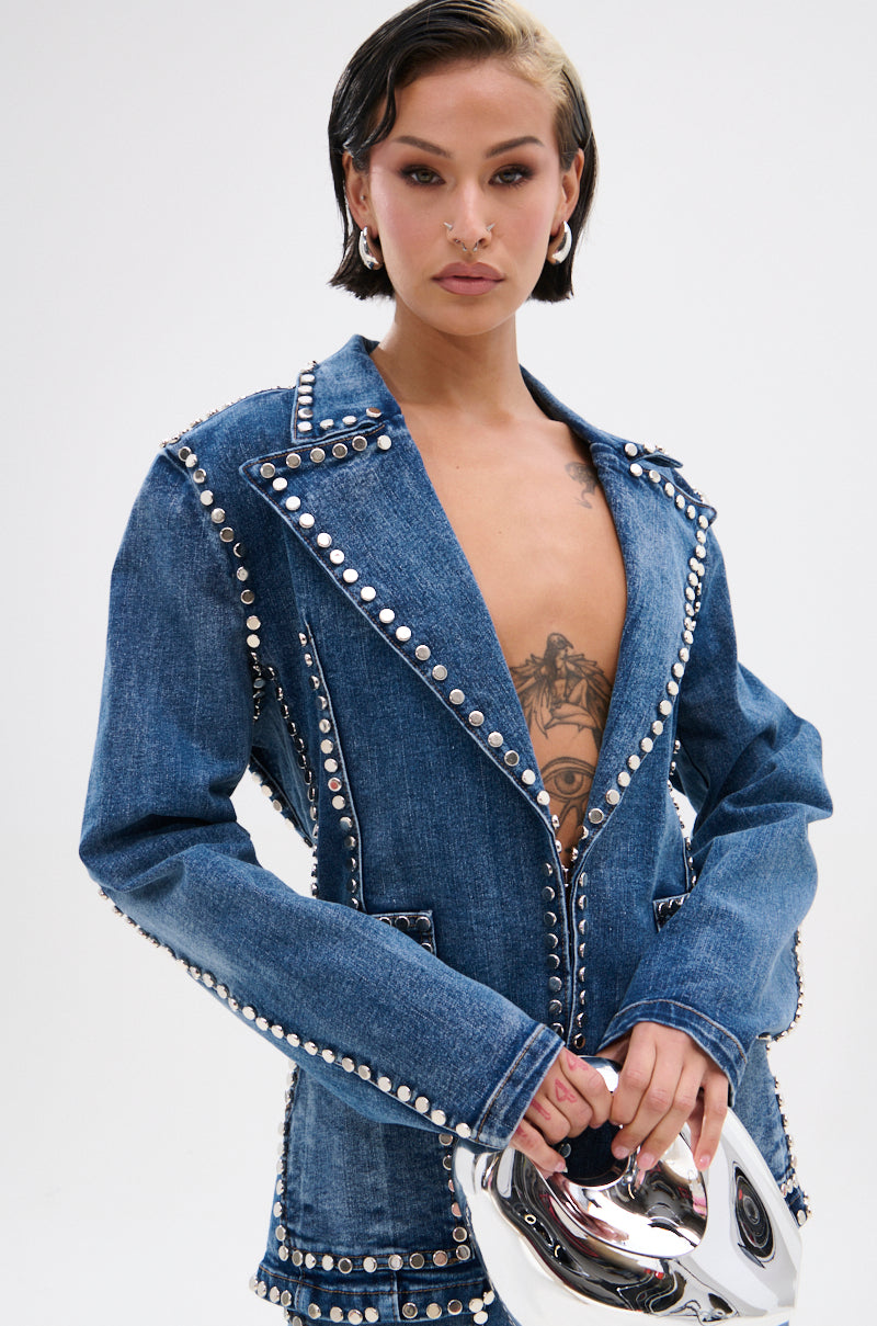A person with short dark hair wears the MY OWN THING STUDDED DENIM BLAZER—tailored fit with a notched lapel and no shirt underneath, revealing a chest tattoo. They hold a shiny silver mask against a light plain background.