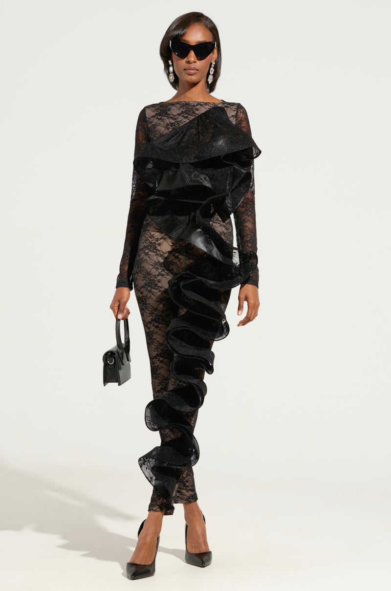 Confidently walking, a person stuns in a RUFFLED LACE CATSUIT-BLACK, paired effortlessly with a small black handbag and sleek black pointed heels. Their look is completed with chic sunglasses against a plain, light-colored background.