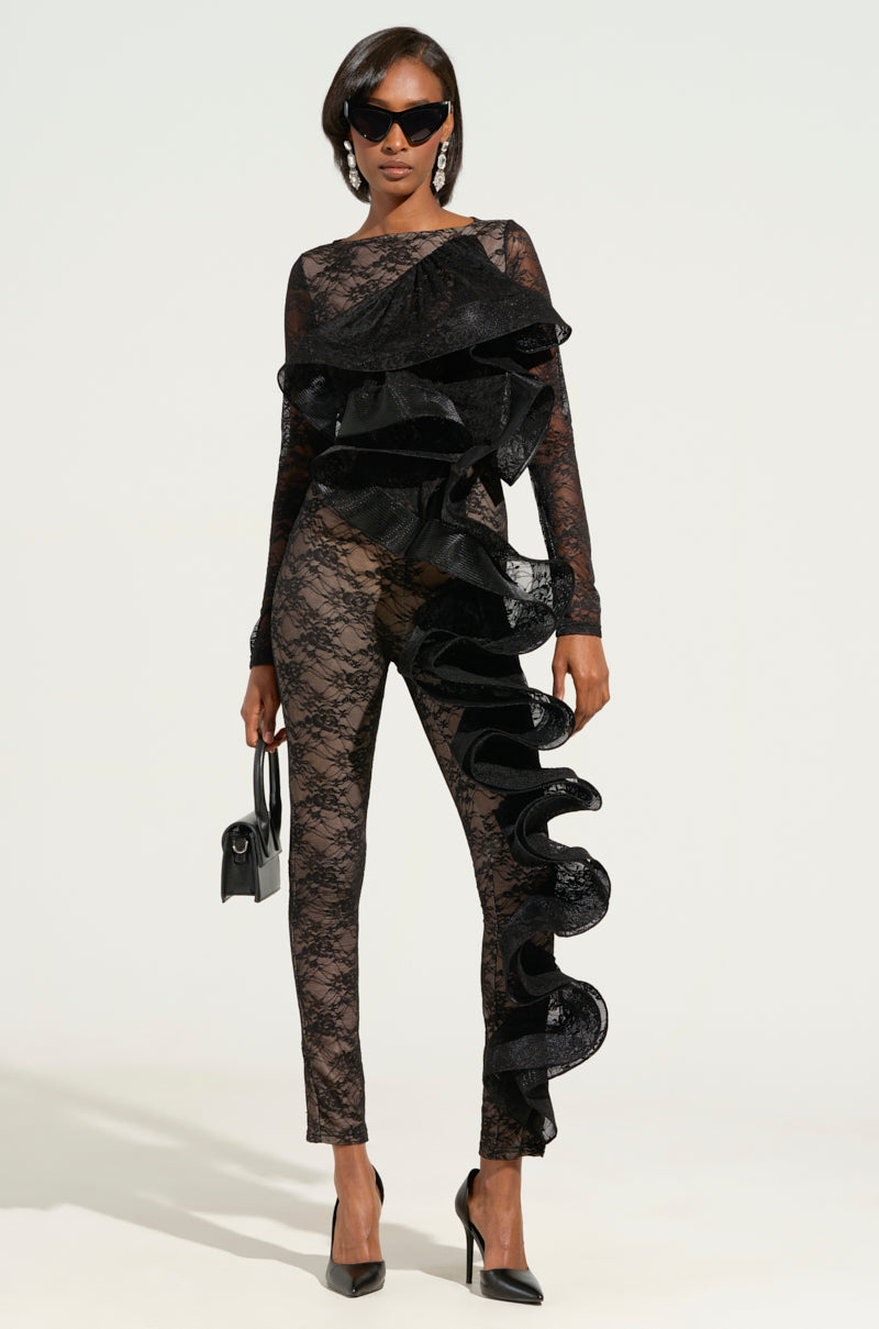 Clad in the RUFFLED LACE CATSUIT-BLACK, a person showcases its elegant ruffle accents along the front and side. They finish the ensemble with black sunglasses, heels, and a small handbag, set against a plain background.