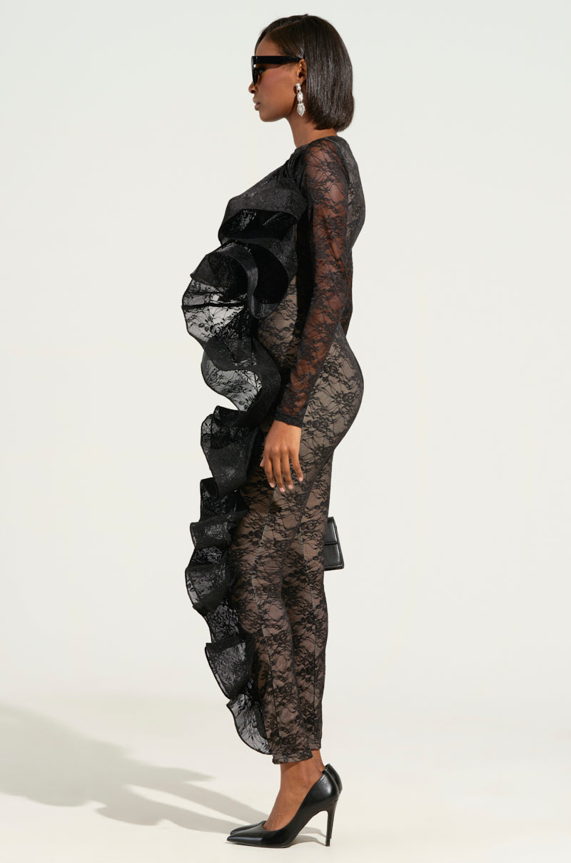 A woman models in profile wearing the RUFFLED LACE CATSUIT-BLACK, showcasing its sleek, contour-defining design with delicate ruffle accents. She completes her ensemble with dark sunglasses, earrings, and black high heels against a light, neutral backdrop.