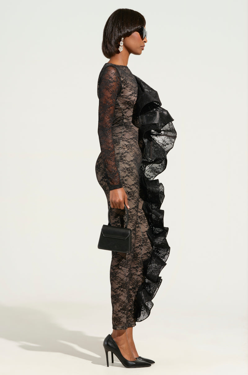 A person is posing in profile, wearing the RUFFLED LACE CATSUIT-BLACK featuring exquisite lace detailing and ruffle accents. High heels elevate the look, while a small black handbag adds charm. They sport sunglasses and earrings against a plain background.