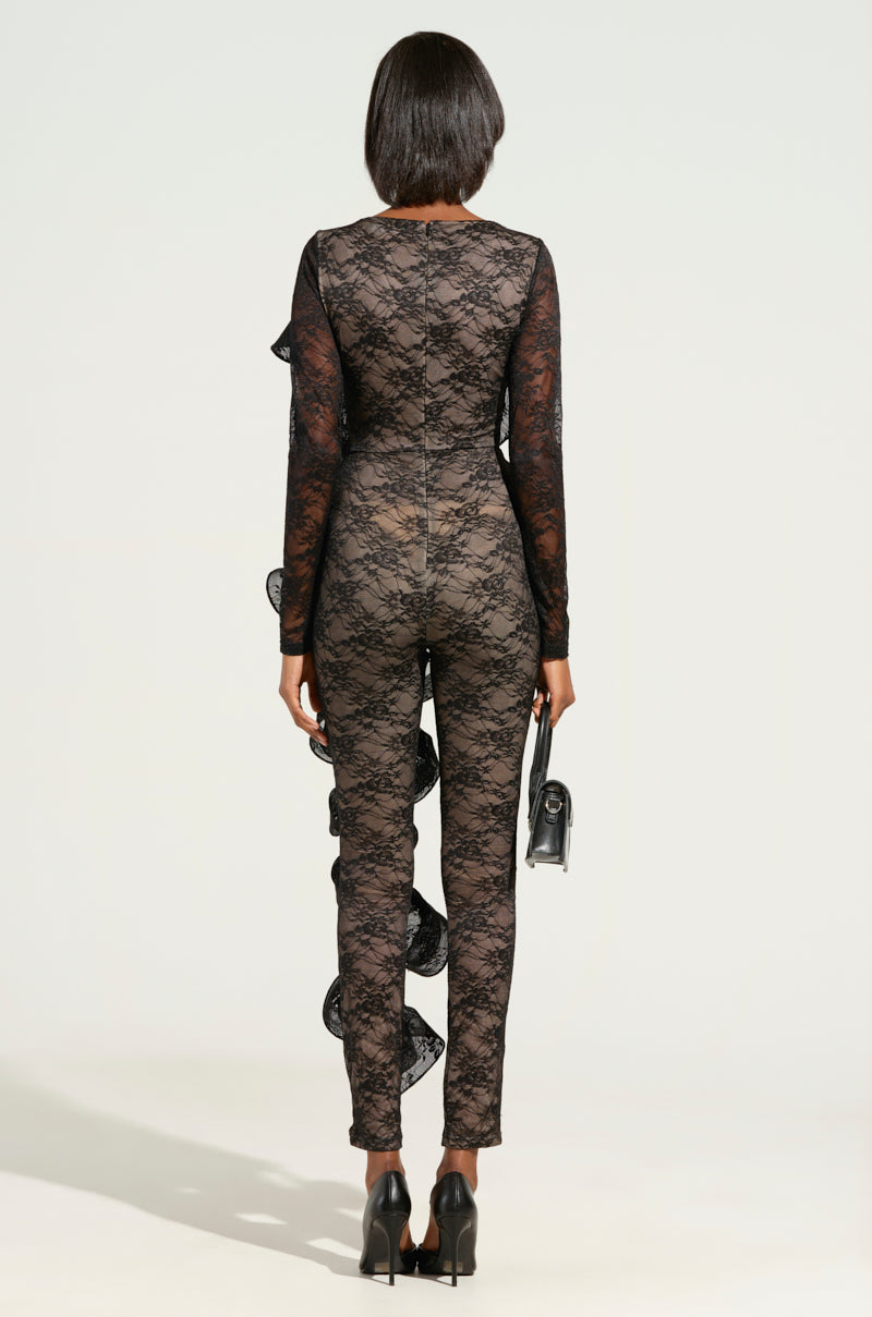 The individual faces away from the camera, donned in the RUFFLED LACE CATSUIT-BLACK, which showcases sophisticated ruffle details along the sides. They hold a petite purse in their left hand and finish off the look with black high heels against a simple, light-colored background.