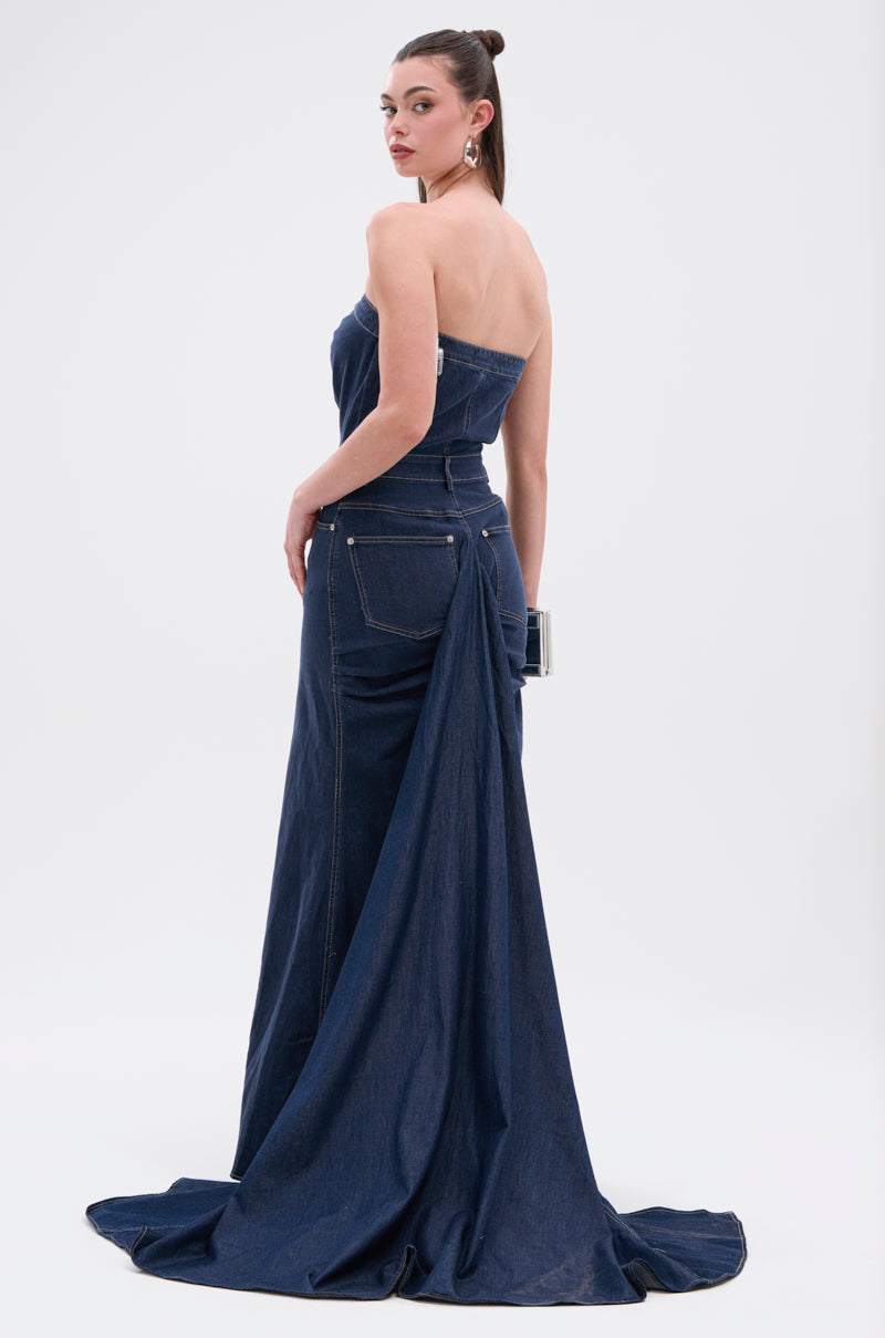 A woman stands facing away against a white background, wearing the LUNA DENIM MAXI DRESS—a dark-wash, strapless maxi with a floor-length train and back pockets. She carries a clutch and her hair is styled in a high, sleek bun.