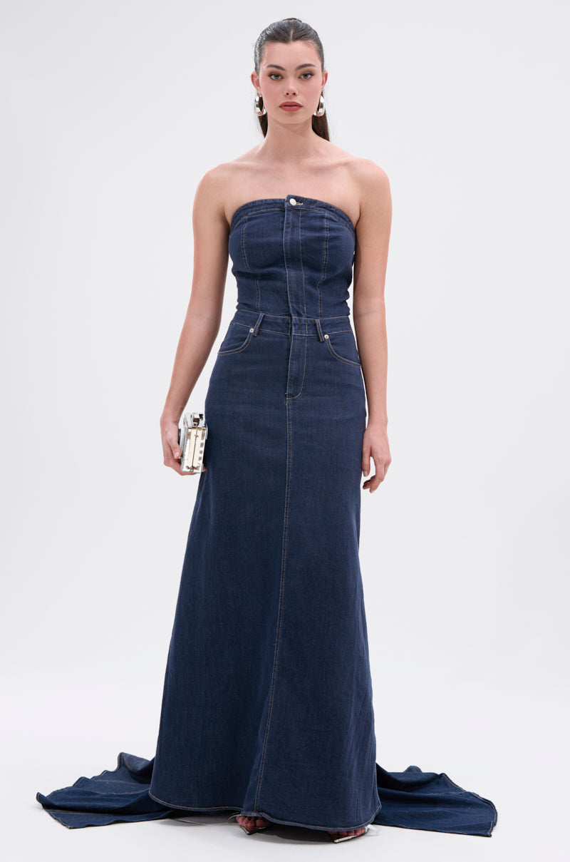 A woman stands against a plain background in the LUNA DENIM MAXI DRESS, featuring a dark-wash, strapless neckline, visible seams, and a small train. She holds a metallic clutch and has her hair pulled back.