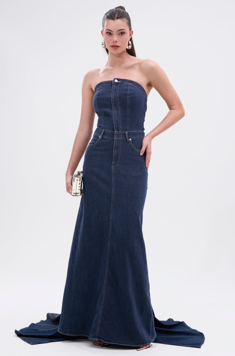 A woman stands against a plain white background, holding a small handbag and wearing the LUNA DENIM MAXI DRESS—a dark-wash, strapless maxi dress with a fitted bodice.