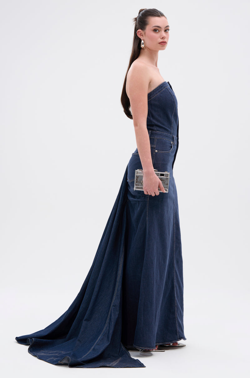 A woman stands in profile against a white background, wearing the LUNA DENIM MAXI DRESS with a strapless neckline and long train. She holds a small clutch; her straight brown hair and earrings complete the look.