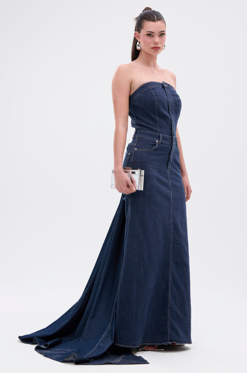 A woman poses against a plain background in the LUNA DENIM MAXI DRESS, featuring a strapless neckline, dark-wash denim, and a small train. She accessorizes with a silver clutch, large hoop earrings, and a sleek ponytail.