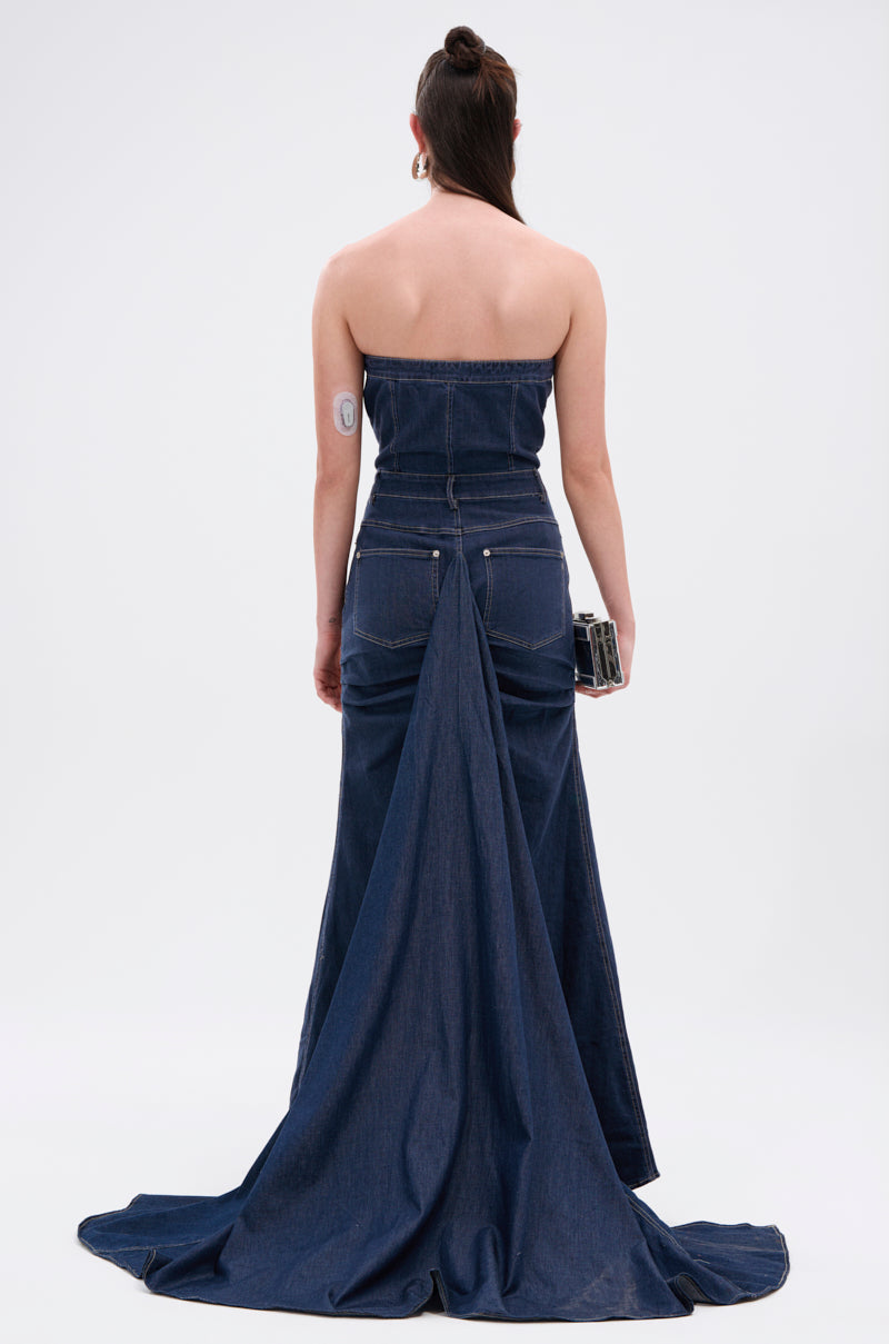 A person in the LUNA DENIM MAXI DRESS featuring a strapless neckline, dark-wash denim, and dramatic train stands facing away on a plain white background, holding a clutch with long brown hair styled up to reveal an arm patch.