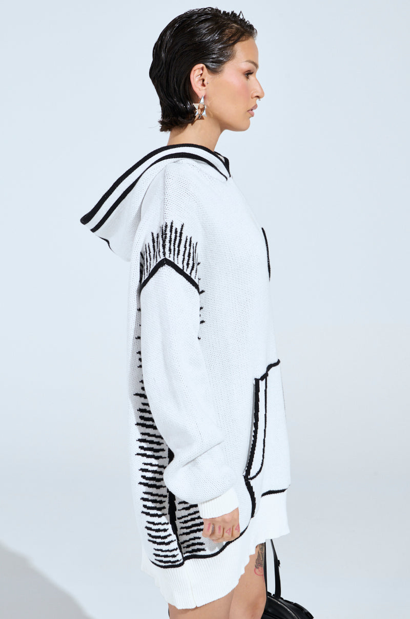 A person with short wet hair wears the SKETCH IT KNIT SWEATSHIRT DRESS, featuring black line patterns and artsy trim. They accessorize with earrings and carry a black handbag against a plain white background.