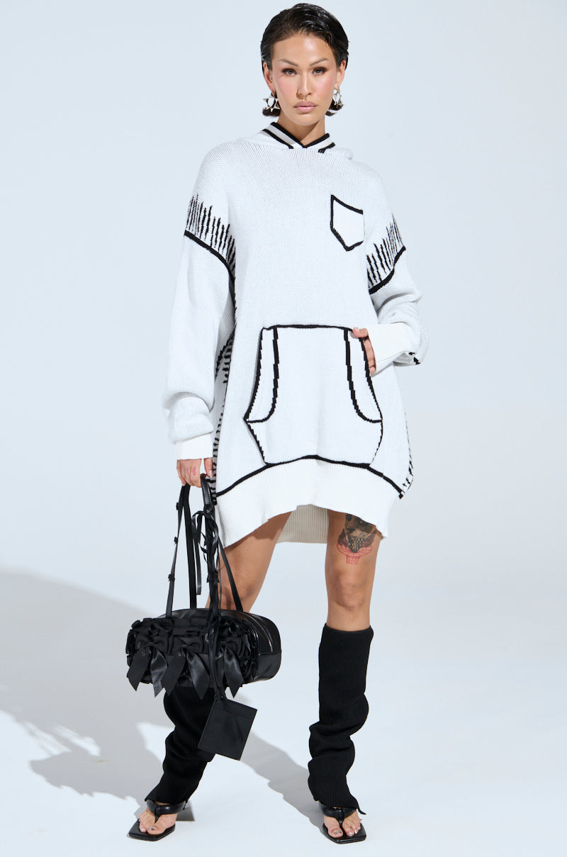 A person exudes confidence in the SKETCH IT KNIT SWEATSHIRT DRESS with artsy trim and a front pocket, holding a black handbag. They complement their look with black knee-high leg warmers over flip-flops and have their hair styled back.