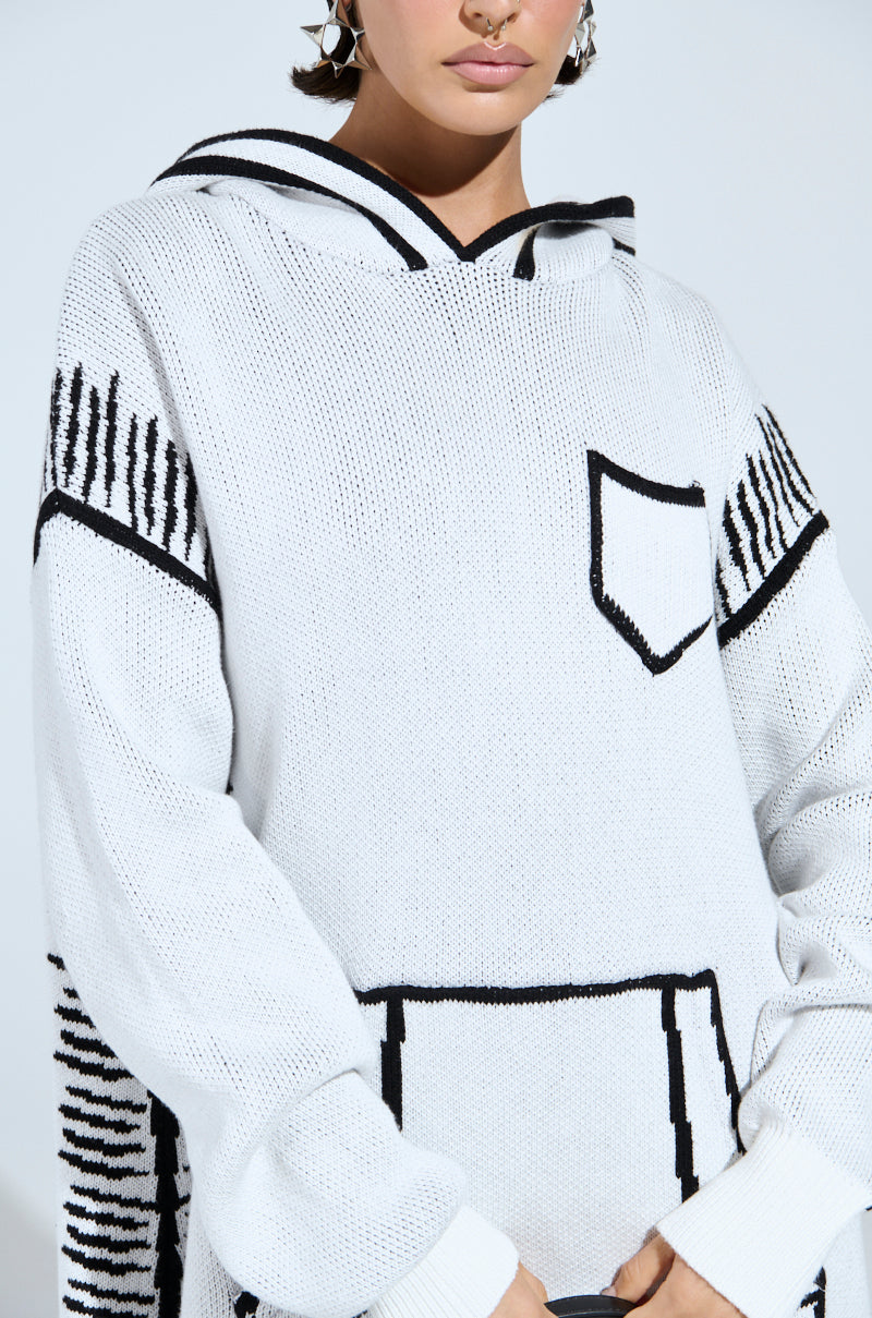 The individual wears the SKETCH IT KNIT SWEATSHIRT DRESS, featuring a white hooded design with black artsy trim details, side stripes, and a front pocket. Hoop earrings and a septum piercing complete their edgy look as they stand against a light background.