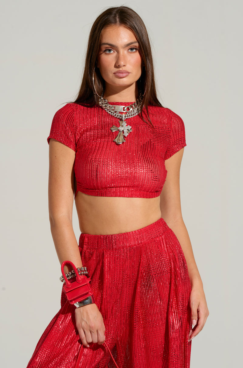 A person with long brown hair is wearing the AKIRA Label's JUST LIKE MAGIC METALLIC OPEN BACK TOP IN RED, paired with a matching skirt. They accessorize with large hoop earrings, a chunky silver necklace adorned with pendants, and a red bracelet. The plain, light-colored background accentuates their cropped silhouette.