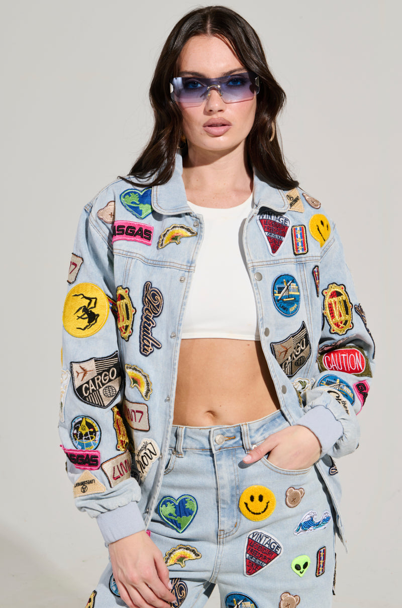 A woman wearing the AUSTIN PATCH DENIM JACKET, embellished with vibrant embroidered patches, pairs it with jeans and stands confidently. Her long brown hair frames her large sunglasses and white crop top against a neutral background.