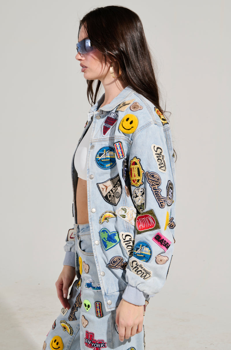 A woman confidently poses in the AUSTIN PATCH DENIM JACKET, a stylish piece of outerwear featuring light blue denim with various colorful embroidered patches and badges. Paired with matching pants, a white top, and sunglasses, she stands against a plain background.