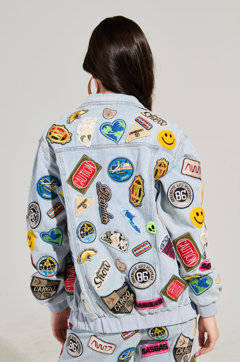 A person with long, dark hair is seen from behind wearing the AUSTIN PATCH DENIM JACKET, a light blue denim piece adorned with colorful embroidered patches featuring smiley faces, planets, and various logos. They stand against a plain background.