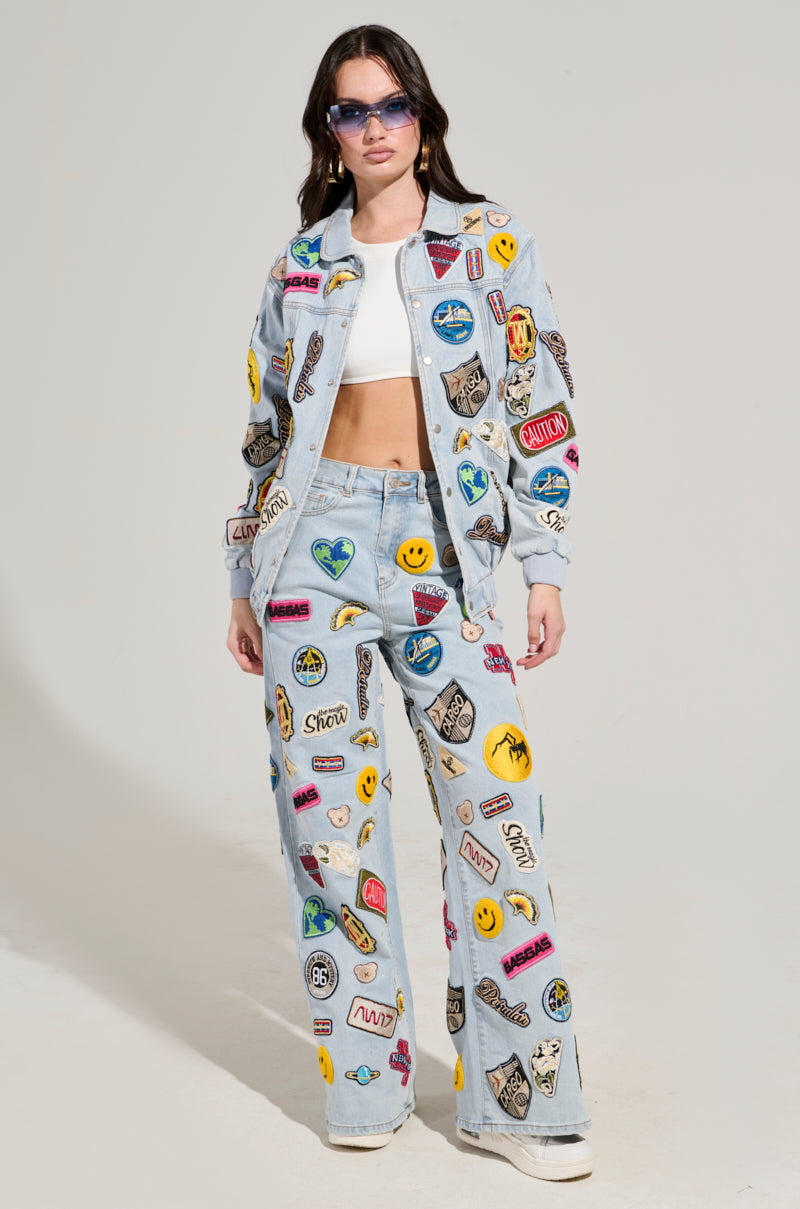 A person is wearing the AUSTIN PATCH DENIM JACKET, embellished with embroidered patches and designs such as smiley faces and assorted logos. They have long hair and sunglasses, paired with white sneakers and a white crop top, posed against a plain background. The vibrant jacket stands out in its uniqueness.