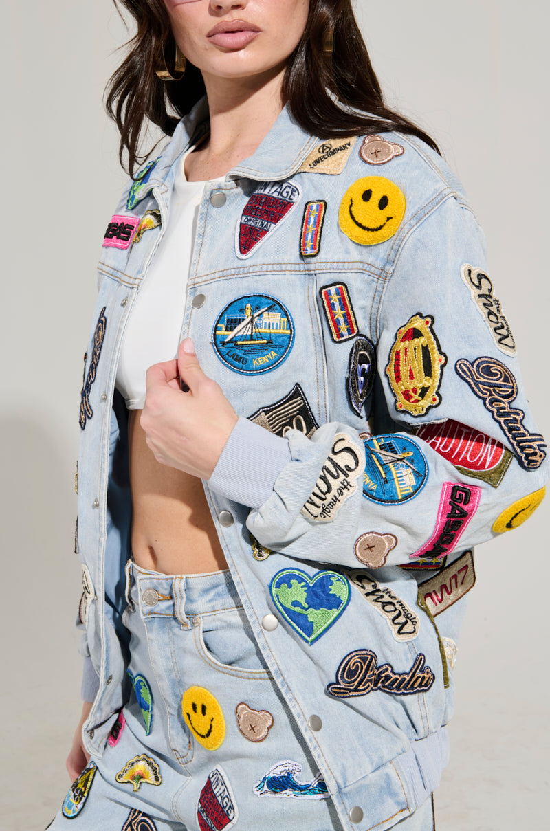 A person is wearing the AUSTIN PATCH DENIM JACKET, featuring colorful embroidered patches such as smiley faces, hearts, and logos, paired with a matching skirt. They have long hair and are posing in a neutral setting.