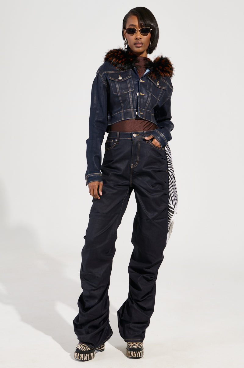 A person stands confidently in a Y2K-inspired outfit, flaunting the SITTING PRETTY FUR TRIM COATED DENIM JACKET featuring a faux fur trim collar, high-waisted black pants, and zebra-patterned shoes. They sport sunglasses and have a patterned scarf hanging from their waist.
