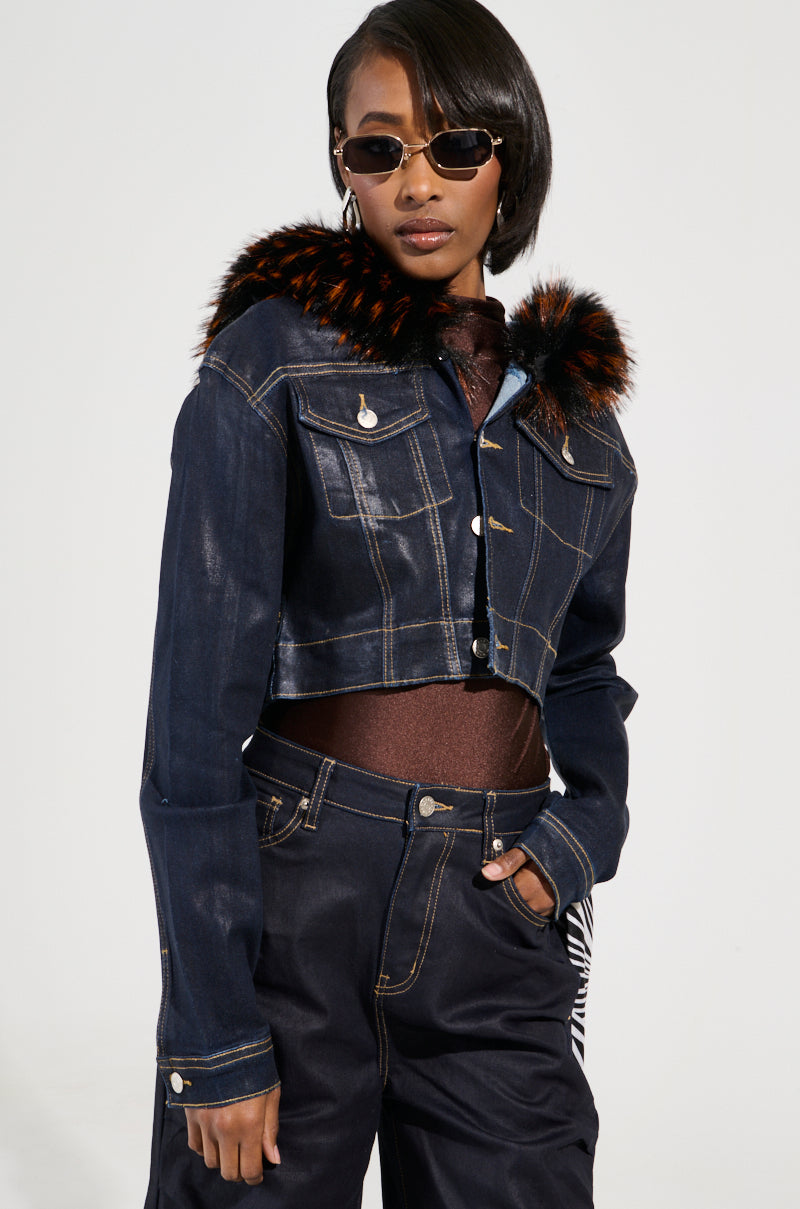 A person confidently poses in the stylish and modern SITTING PRETTY FUR TRIM COATED DENIM JACKET, which is Y2K-inspired and features intricate stitching details. The cropped jacket has a faux fur trim with a brown collar and black tips, paired with dark pants and sunglasses.