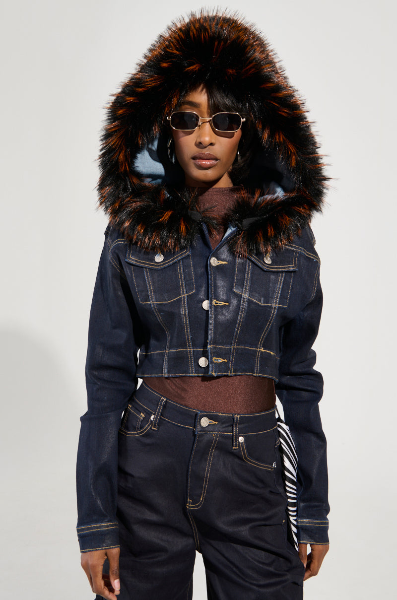 A person stands confidently, sporting the SITTING PRETTY FUR TRIM COATED DENIM JACKET featuring a faux fur trim hood and sunglasses. The black and orange fur design on the hood adds an edgy touch, perfectly complementing the dark jeans for a stylish blend of casual and bold fashion elements.