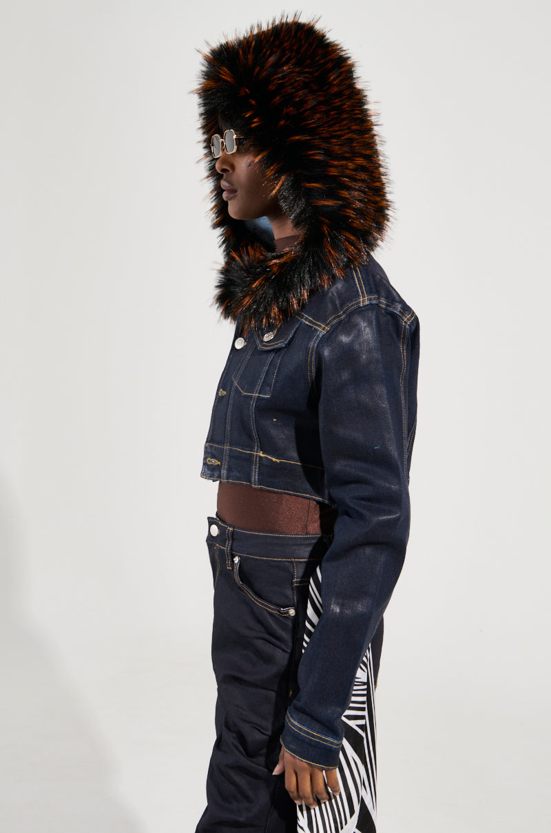 A person is standing in profile against a neutral background, wearing the stylish SITTING PRETTY FUR TRIM COATED DENIM JACKET with a faux fur trim hood and fashionable sunglasses. The Y2K-inspired jacket is cropped and paired with pants that feature a monochrome pattern.