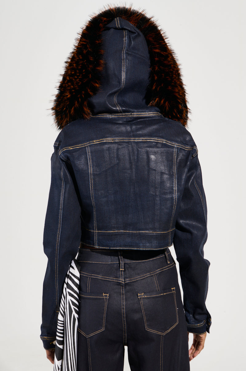 The back view of someone wearing the SITTING PRETTY FUR TRIM COATED DENIM JACKET, a Y2K-inspired dark denim piece with a faux fur trim hood. This cropped jacket complements the dark jeans that have visible seams and feature a black and white striped fabric detail hanging from the side.