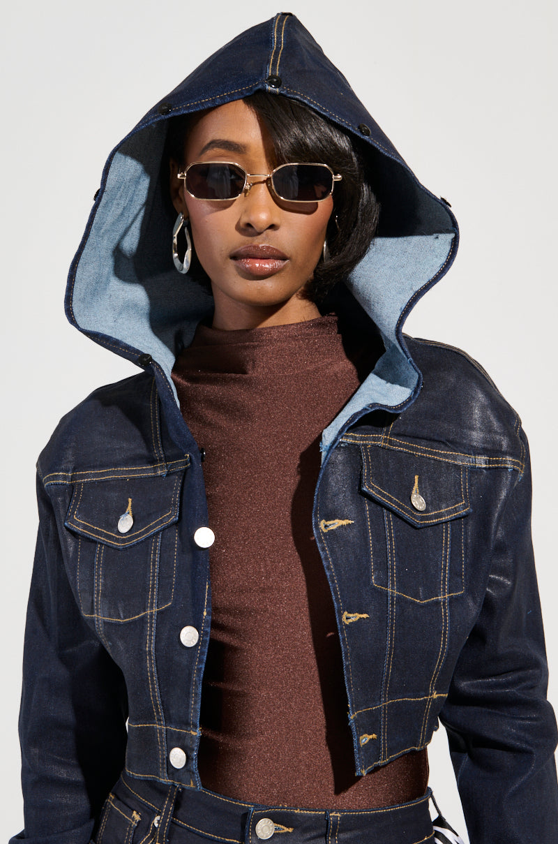 A person wearing sunglasses, hoop earrings, and the Sitting Pretty Fur Trim Coated Denim Jacket, showcasing a Y2K-inspired style with prominent stitching and metallic buttons over a brown top. The jacket features a hood lined with lighter blue fabric, adding a nostalgic twist as the individual looks directly at the camera.
