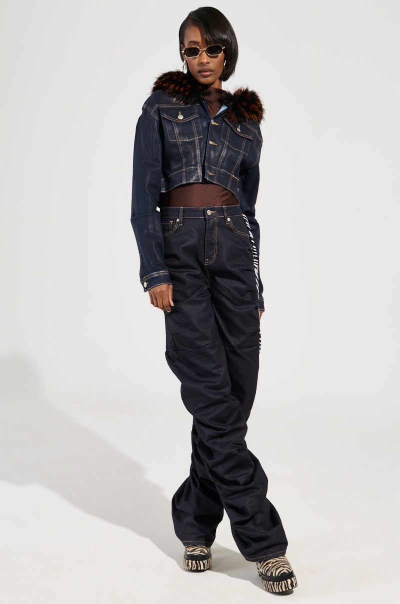 A person wearing sunglasses poses confidently in a Y2K-inspired ensemble. They sport the SITTING PRETTY FUR TRIM COATED DENIM JACKET with wide-leg pants, perfectly complemented by patterned boots. The background is plain and white.