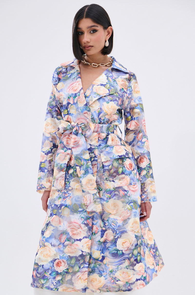 A woman stands before a white backdrop wearing the PLANT THE SEED SHEER TRENCH—a belted, pastel floral organza coat. She has straight dark hair, a statement necklace, and exudes confidence.