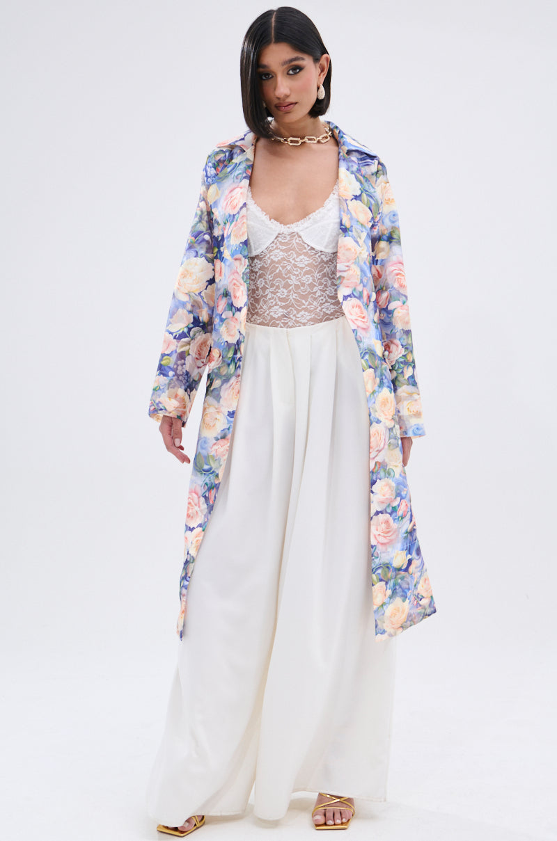 A woman wears the PLANT THE SEED SHEER TRENCH in floral organza over a white lace top and flowing pants, styled with sleek dark hair, gold earrings, a chunky gold necklace, and gold sandals against a plain white background.