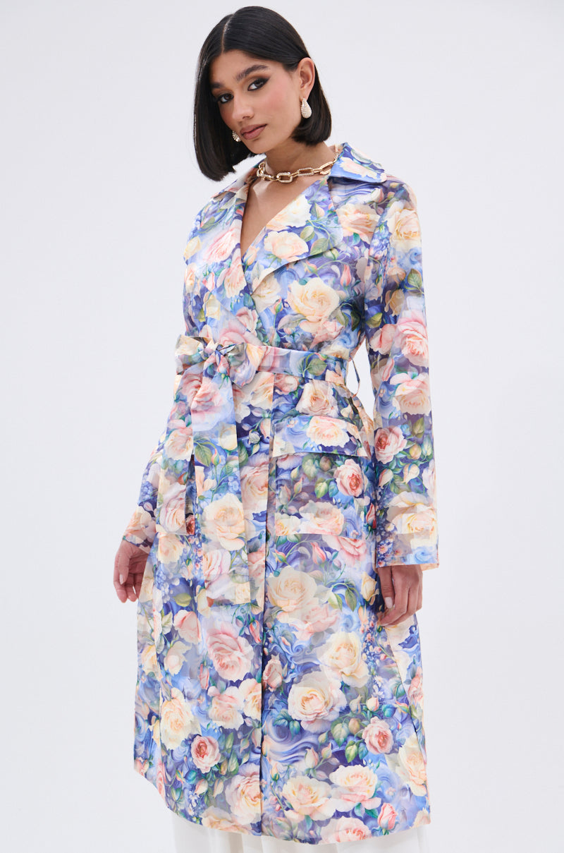 A woman with straight dark hair wears the PLANT THE SEED SHEER TRENCH—a knee-length, belted coat with a vibrant pastel floral print—standing confidently against a plain white background.