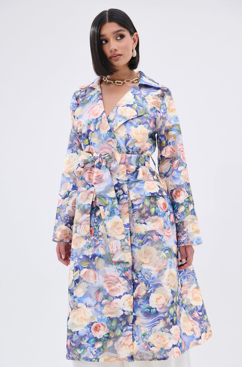 A person wearing the PLANT THE SEED SHEER TRENCH, a floral print organza coat with a matching belt, gold chain necklace, and straight shoulder-length hair stands against a plain white background.