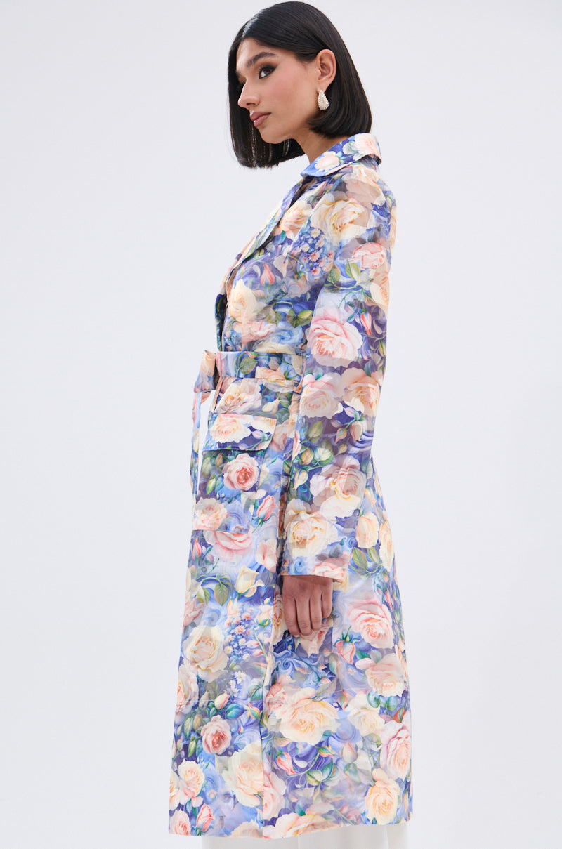 A woman with straight, shoulder-length dark hair wears the PLANT THE SEED SHEER TRENCH—a soft floral print coat—standing in profile against a plain light background.
