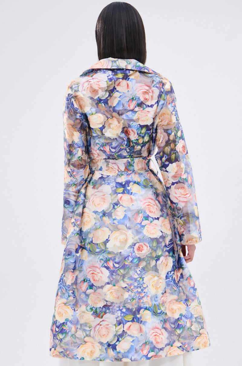 A person with straight, dark hair faces away, wearing the PLANT THE SEED SHEER TRENCH—an organza trench coat with a delicate floral print in pastel pink, blue, and cream on a light background.