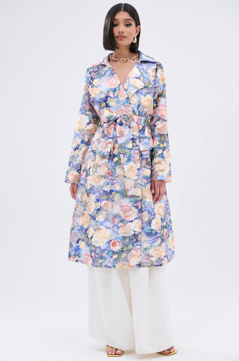 A woman stands against a plain white background wearing the PLANT THE SEED SHEER TRENCH, a long, floral print sheer trench coat with a tie belt, styled over wide-legged white pants and open-toed sandals.