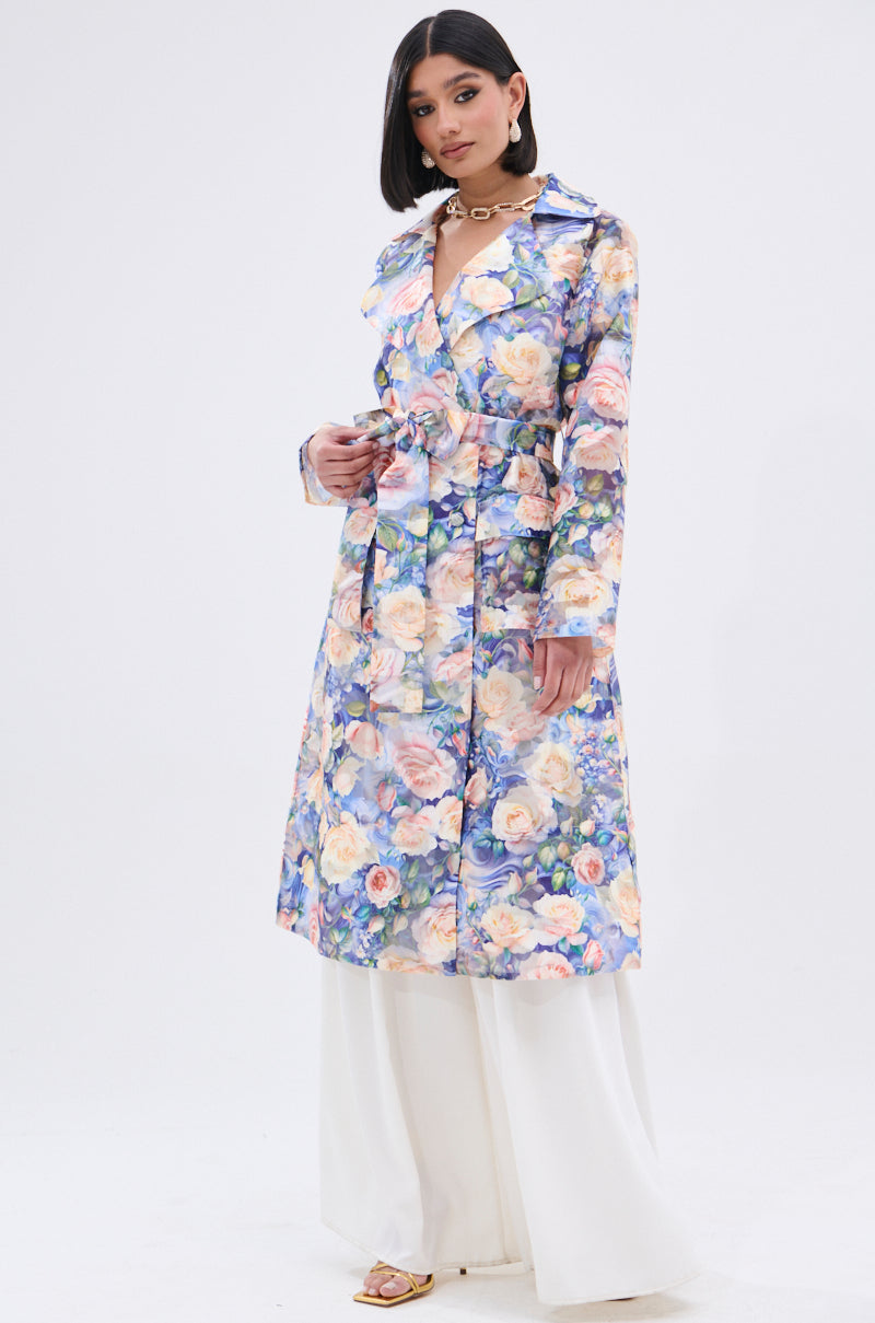 A woman wears the PLANT THE SEED SHEER TRENCH, featuring a colorful floral print, over wide-legged white pants and open-toe sandals against a plain white background.