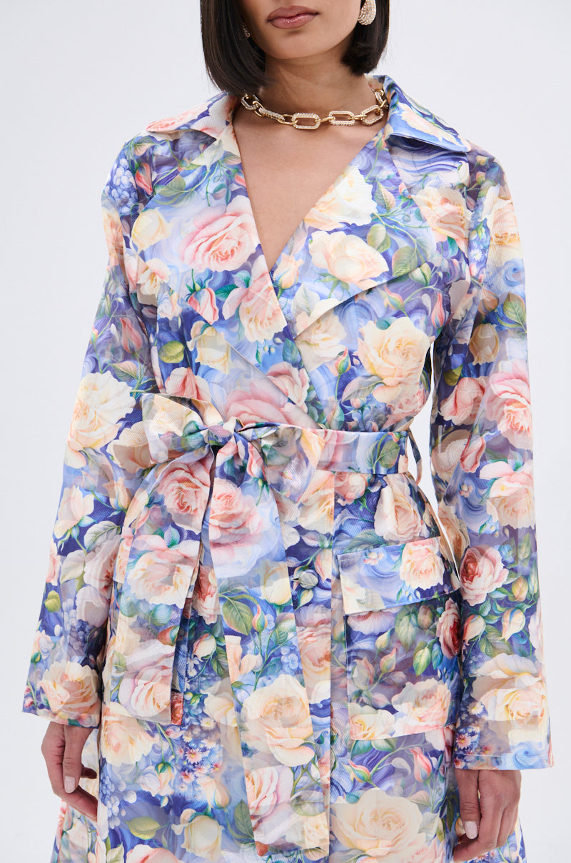 Wearing the PLANT THE SEED SHEER TRENCH, an organza coat with a pink, blue, and yellow floral print, she accents her look with a chunky gold chain necklace while her shoulder-length dark hair frames her face.